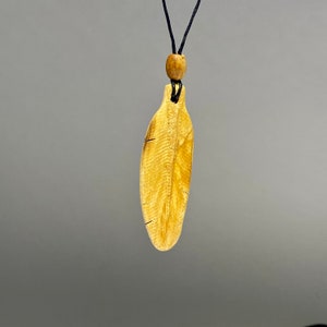 Puede incluir: Un collar con colgante de madera con un colgante en forma de hoja. El colgante es de color marrón claro con vetas de madera visibles y pequeñas grietas. Cuelga de un cordón negro con una cuenta de madera. El fondo es gris liso.