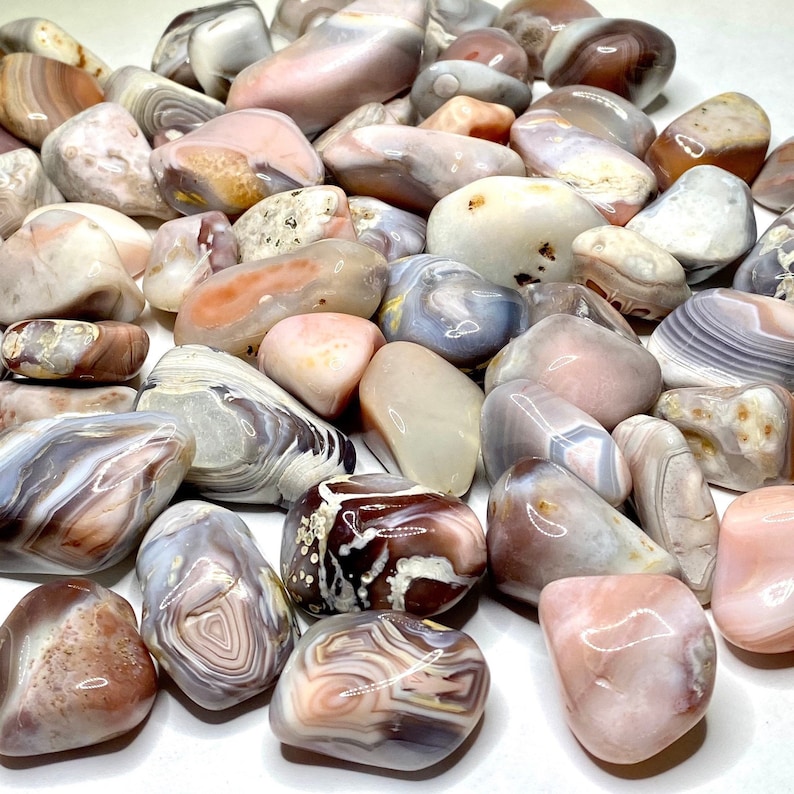 A collection of pink, white, and gray agate tumbled stones from Botswana. The stones have a variety of patterns and textures. The stones are smooth and polished.