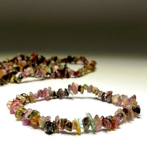A bracelet made of small, chipped, multi-colored gemstones. The bracelet is made of pink, green, brown, black, and yellow gemstones. The bracelet is lying on a white surface.
