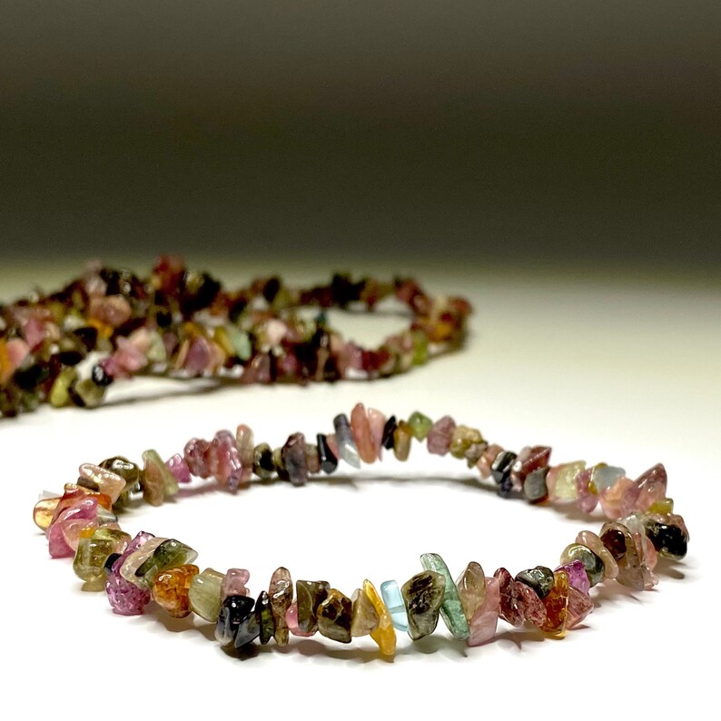 A bracelet made of small, chipped, multi-colored gemstones. The bracelet is made of pink, green, brown, black, and yellow gemstones. The bracelet is lying on a white surface.