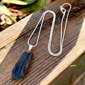 Puede incluir: Un collar de cadena de plata con un colgante de turmalina negra. El colgante es una pieza bruta de turmalina negra sin pulir.