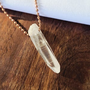 May include: A clear quartz crystal pendant on a rose gold chain. The crystal is long and slender, with a pointed tip. The chain is made of small, round beads.