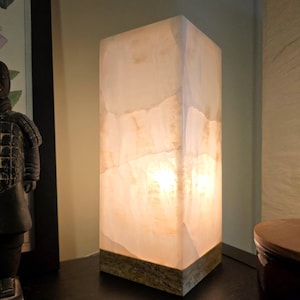 May include: A rectangular table lamp featuring a translucent, off-white stone shade with visible natural veining and warm internal illumination. The lamp is supported by a solid, textured, earth-toned stone base with a mottled, granular appearance.