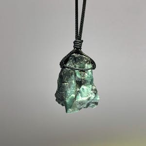 May include: A raw emerald gemstone pendant on a black cord necklace. The emerald is a deep green color and has a rough, natural texture.