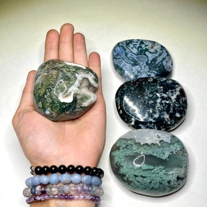 May include: Four moss agate stones, each with a unique pattern of green, black, and white. The stones are smooth and polished, and they are arranged in a row on a white surface.