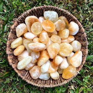 May include: A woven basket filled with polished citrine gemstones. The stones vary in size and shape, displaying shades of yellow, orange, and white. The basket is brown, and the background is green grass.