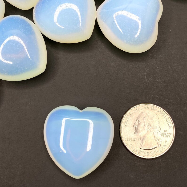 A close-up of a heart-shaped, pale blue opal stone. The stone is smooth and polished. The stone is sitting on a black surface. A US quarter is next to the stone for size comparison. The quarter has the words 'United States of America', 'Liberty', 'In God We Trust', and 'Quarter Dollar' inscribed on it.