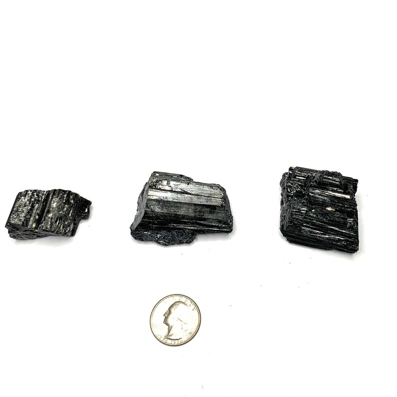 Three black tourmaline crystals of varying sizes and shapes. The crystals are raw and have a natural, rough texture. The crystals are arranged in a row on a white background.