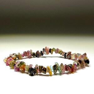 A bracelet made of small, chipped, multi-colored gemstones. The gemstones are pink, green, brown, and black. The bracelet is on a white surface.