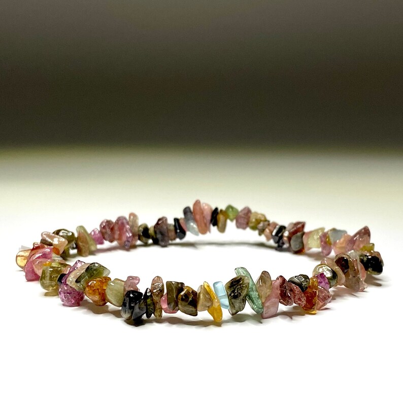 A bracelet made of small, chipped, multi-colored gemstones. The gemstones are pink, green, brown, and black. The bracelet is on a white surface.