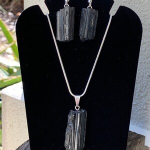 Puede incluir: Un conjunto de collar y pendientes de turmalina negra. El collar tiene una cadena larga de plata con un colgante de turmalina negra. Los pendientes son de plata con piedras de turmalina negra.