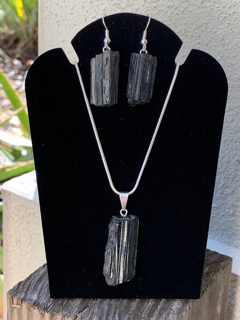 Puede incluir: Un conjunto de collar y pendientes de turmalina negra. El collar tiene una cadena larga de plata con un colgante de turmalina negra. Los pendientes son de plata con piedras de turmalina negra.