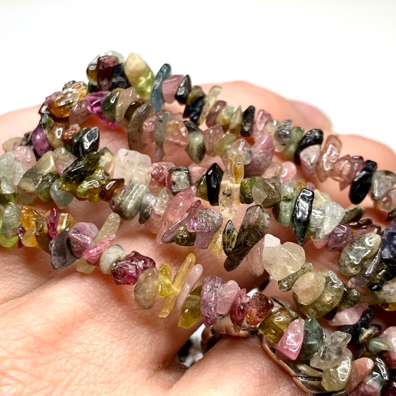 A strand of multi-colored chip gemstone beads. The beads are a mix of pink, green, black, and yellow. The beads are irregular in shape and have a rough, natural texture.