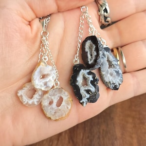 May include: Silver chain necklaces with geode pendants. One necklace features white and tan geodes, while the other has black and gray geodes. The pendants have a natural, crystal-like appearance, with hollow centers.