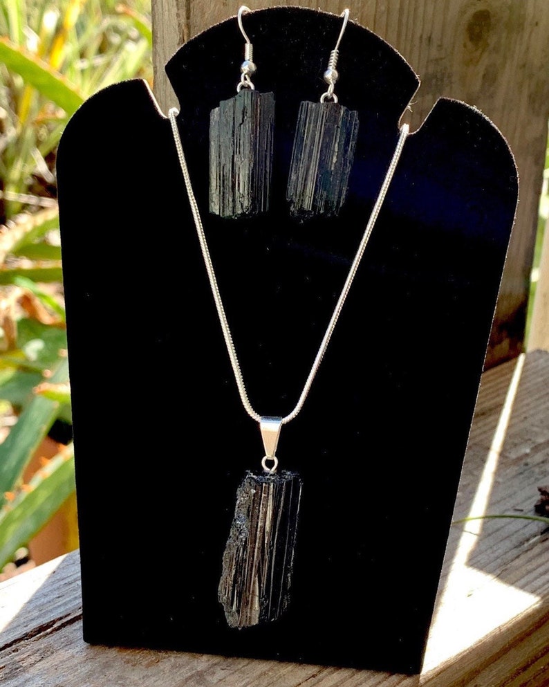 Puede incluir: Un conjunto de collar y pendientes de turmalina negra. El collar tiene una cadena larga de plata con un colgante de turmalina negra. Los pendientes son de plata con piedras de turmalina negra.