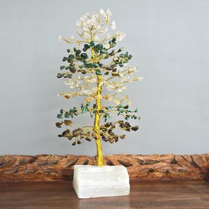 May include: A decorative gemstone tree with a gold-colored wire trunk and branches. The tree is adorned with various colorful gemstones, including clear, green, and brown stones. It is set on a white rectangular base, creating a unique and artistic display.