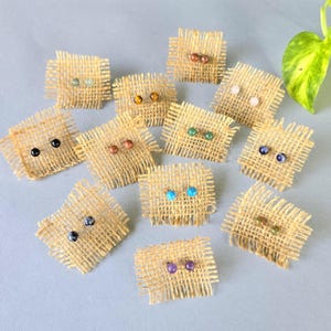 May include: A collection of twelve pairs of small, round, spherical stud earrings in various colors, including black, blue, green, brown, and purple. Each pair is mounted on a small, square piece of light brown, frayed-edge burlap fabric.