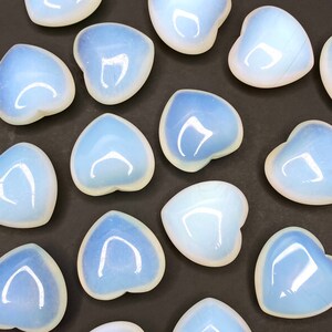A collection of heart shaped polished opalite stones. The stones are a milky white color with a slight blue tint. The stones are arranged in a pattern on a black surface.