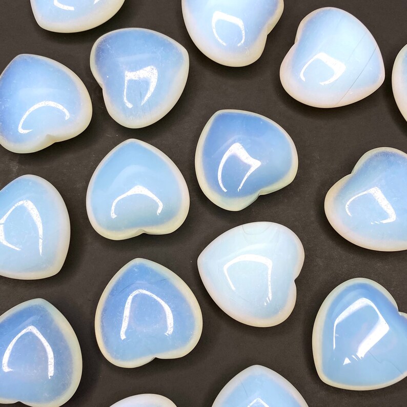 A collection of heart shaped polished opalite stones. The stones are a milky white color with a slight blue tint. The stones are arranged in a pattern on a black surface.