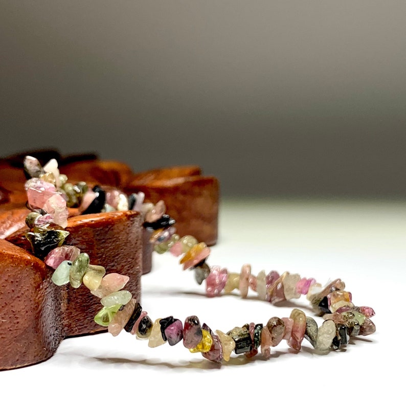A bracelet made of small, multi-colored gemstones. The bracelet is made of pink, green, black, and yellow gemstones. The bracelet is lying on a wooden surface.