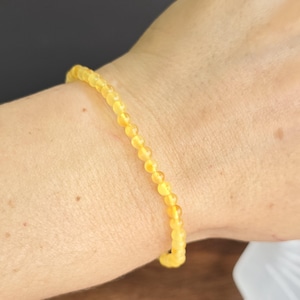 May include: A fine bracelet made of small, round, yellow beads. The bracelet is displayed on a wrist, highlighting its simple and elegant design. The beads have a warm, golden hue, possibly indicating a natural material.