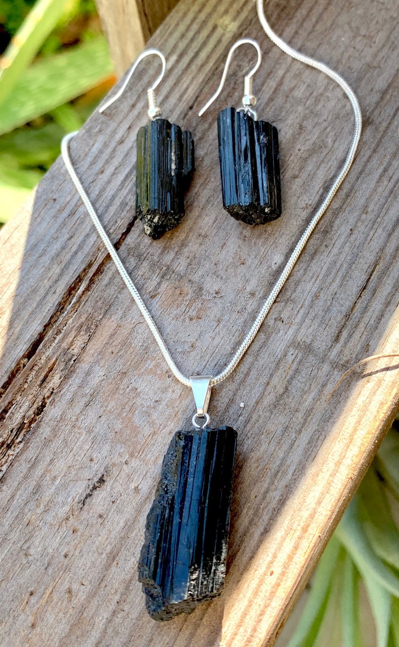 Puede incluir: Un conjunto de collar y pendientes de turmalina negra. El collar tiene una cadena larga de plata con un colgante de turmalina negra. Los pendientes son de plata con piedras de turmalina negra.