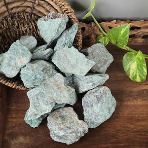 May include: A collection of raw, unpolished green stones with rough, jagged textures and earthy mineral variations. The stones are displayed in a woven brown basket, showcasing natural shades of sage and forest green with hints of brown.