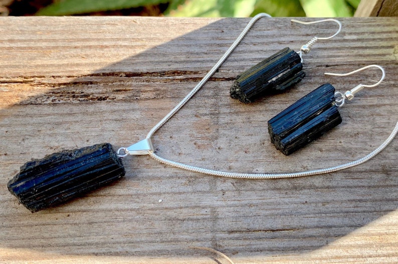 Puede incluir: Un conjunto de collar y pendientes de turmalina negra con cadenas de plata. El collar presenta un gran colgante de turmalina negra en bruto. Los pendientes son simples aros de plata con peque&ntilde;as piedras de turmalina negra en bruto.