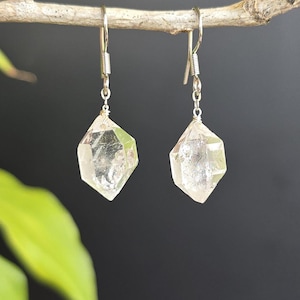 Puede incluir: Un par de pendientes de cristal de cuarzo transparente. Cada pendiente presenta un cristal facetado en forma de diamante suspendido de un gancho plateado. Los cristales son transparentes con estructuras internas visibles, sobre un fondo oscuro.