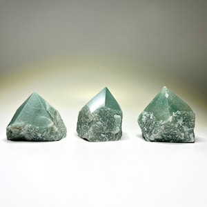 May include: Three green aventurine crystal points, each with a rough, natural texture. The points are arranged in a row on a white surface.