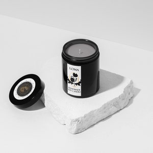 Puede incluir: Un tarro de vela negro con una etiqueta blanca que dice "LOMA HANDMADE SOY CANDLE". La vela est&aacute; sobre una roca blanca y la tapa est&aacute; a un lado.