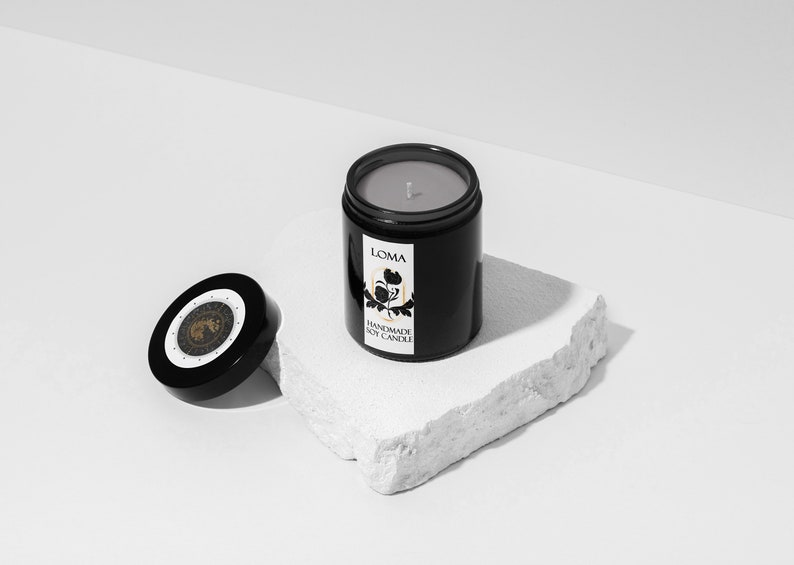 Puede incluir: Un tarro de vela negro con una etiqueta blanca que dice "LOMA HANDMADE SOY CANDLE". La vela est&aacute; sobre una roca blanca y la tapa est&aacute; a un lado.