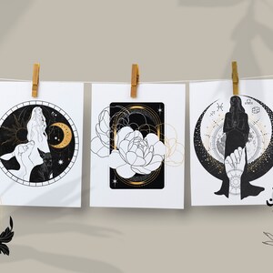 Puede incluir: Tres ilustraciones en blanco y negro con detalles dorados sobre papel blanco, colgadas de una cuerda para tender la ropa con pinzas de madera. Las ilustraciones representan a una mujer con una luna creciente, una flor y una mujer con una luna creciente y un ciervo.