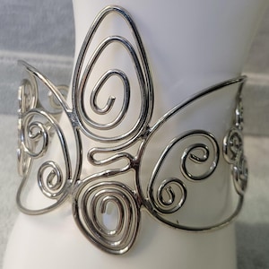 May include: A silver-toned cuff bracelet with an openwork design. The bracelet features a large, central butterfly-like shape with swirling wire details. The open design allows the skin to show through, creating a delicate and airy look.