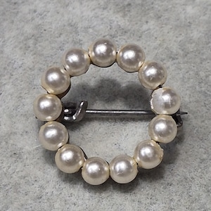 May include: A small, round brooch featuring a ring of cream-colored faux pearls. The pearls are evenly spaced and attached to a silver-toned metal pin backing. The brooch is a classic jewelry piece.