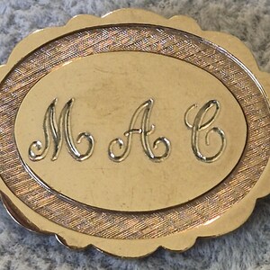 May include: Gold-toned oval brooch with scalloped edges. The center features an oval with the engraved initials "M A C" in a script font. The outer border has a textured design.
