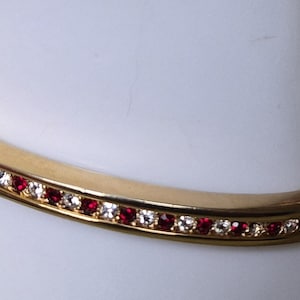 May include: A gold-colored bangle bracelet with a row of alternating red and clear gemstones. The bracelet is curved and sits on a white surface. The gemstones are small and evenly spaced.