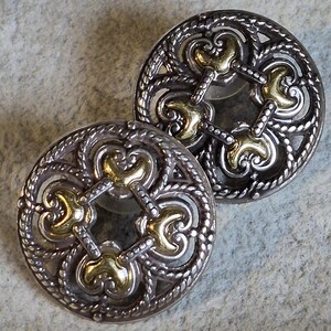 May include: Two ornate, silver-toned buttons with gold-colored accents. The buttons feature a detailed, openwork design with a central pattern and a rope-like border. The gold accents are in a quatrefoil shape.