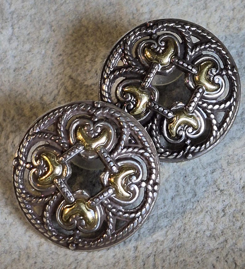 May include: Two ornate, silver-toned buttons with gold-colored accents. The buttons feature a detailed, openwork design with a central pattern and a rope-like border. The gold accents are in a quatrefoil shape.