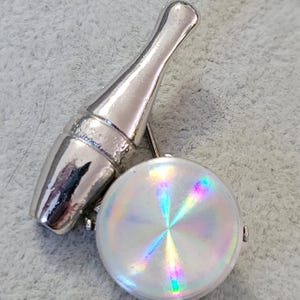 May include: A silver-toned brooch featuring a champagne bottle and a round, iridescent disc. The bottle has a detailed design, and the disc reflects a spectrum of colors. The brooch is likely made of metal.