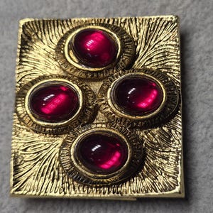 Peut inclure: Broche rectangulaire dorée ornée de quatre pierres précieuses ovales rouge foncé. La broche présente une surface texturée et gravée avec un motif floral ou en forme de feuille. Les pierres sont serties dans des chatons circulaires dorés.