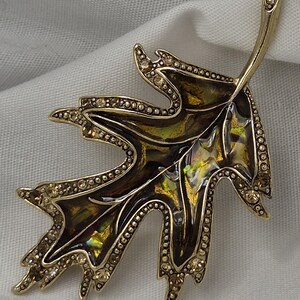May include: A gold-coloured leaf-shaped brooch with an intricate design. The brooch has a dark brown and iridescent green centre, outlined with small, sparkling crystals. The edges are also decorated with crystals.