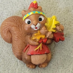May include: A brown squirrel figurine wearing a red and yellow headdress and holding a bunch of yellow and red leaves.