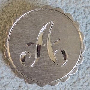 May include: A silver-colored, circular monogram with a scalloped edge. The letter "A" is prominently displayed in a cursive font within the circle. The surface has a brushed metal texture.