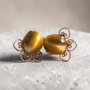 May include: A pair of gold-toned earrings featuring oval-shaped, chatoyant tiger's eye gemstones. Each stone is set within a delicate, openwork metal frame composed of intricate, heart-shaped filigree loops that surround the central cabochon.