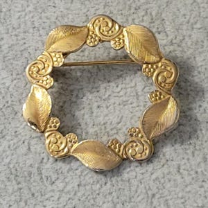 May include: A gold-toned circular brooch with a leaf and scroll design. The brooch features a pin closure on the back. The leaves are detailed with etched veins, and the design is symmetrical. The brooch is likely vintage or antique.