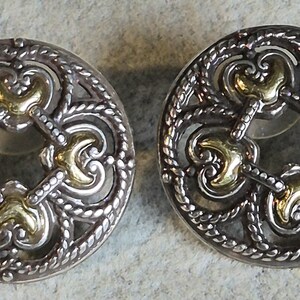 May include: A pair of silver-toned stud earrings with gold-colored accents. The earrings have a circular design with an openwork pattern featuring ornate swirls and a rope-like border. The gold accents are in the center of the design.