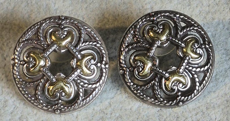May include: A pair of silver-toned stud earrings with gold-colored accents. The earrings have a circular design with an openwork pattern featuring ornate swirls and a rope-like border. The gold accents are in the center of the design.