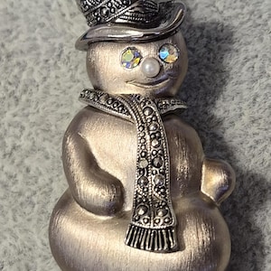May include: Silver-toned snowman brooch with a top hat and scarf. The snowman has crystal eyes and a pearl nose. The scarf and hat are detailed with small, round embellishments. A festive winter accessory.