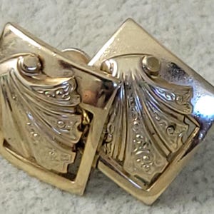 May include: A pair of gold-toned square cufflinks. Each cufflink features an ornate, symmetrical design with a central, stylized fan shape and delicate scrollwork. The cufflinks are connected by a small, hinged bar.